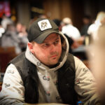 Ein professioneller Pokerspieler mit Sonnenbrille und Kappe sitzt an einem Spieltisch und betrachtet konzentriert seine Karten. Um ihn herum sind Chips und Karten verstreut.