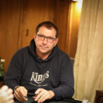Ein professioneller Pokerspieler in einem dunklen Anzug sitzt an einem Pokertisch und betrachtet konzentriert seine Karten. Im Hintergrund sind andere Spieler und Chips zu sehen.