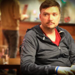 Ein Mann in einem dunkelblauen Anzug und einer Krawatte spielt konzentriert Poker an einem grünen Pokertisch in einem eleganten Casino. Er hält Karten in der Hand und betrachtet mit ern