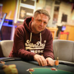 Ein professioneller Pokerspieler mit Sonnenbrille und Kappe sitzt an einem Pokertisch und betrachtet konzentriert seine Karten. Im Hintergrund sind weitere Spieler und Chips zu sehen.