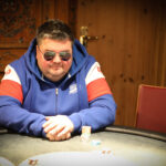 Ein Mann in einem dunkelblauen Anzug und einer Krawatte steht vor einer Pokertisch mit Spielkarten und Pokerchips. Er hält eine Hand mit Karten und schaut mit einem ernsten Gesichtsausdr