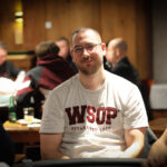 Ein Mann in einem dunkelblauen Anzug und einer roten Krawatte sitzt an einem Pokertisch und betrachtet konzentriert seine Karten. In der Mitte des Tisches liegen Chips und Spielkarten