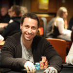 Ein Mann sitzt an einem Pokertisch und betrachtet konzentriert seine Karten. In der Hand hält er Pokerchips und neben ihm liegen Spielkarten auf dem Tisch. Im Hintergrund sind weitere Spieler