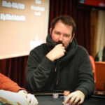 Ein professioneller Pokertisch mit grünem Filz, umgeben von Stühlen und Chips. Drei Spieler sitzen mit ernstem Gesichtsausdruck an ihren Plätzen, während sie Karten halten und auf ihre