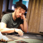 Ein Mann in einem schwarzen Anzug und einer weißen Hemd steht vor einem Pokertisch, auf dem Karten und Pokerchips liegen. Er betrachtet konzentriert seine Karten und wirkt entschlossen.