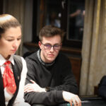 Ein Mann in einem dunkelblauen Anzug sitzt an einem Pokertisch und hält zwei Spielkarten in der Hand. Er schaut konzentriert auf den Tisch, auf dem Pokerchips und Spielkarten verte