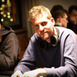 Ein Mann in einem dunklen Anzug sitzt an einem Pokertisch und hält Karten in der Hand. Im Hintergrund sind andere Spieler und Pokerchips zu sehen.