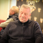 Ein Mann in einem schwarzen Anzug und einer schwarzen Sonnenbrille spielt Poker an einem Tisch in einem eleganten Casino. Er hält Karten in der Hand und setzt Pokerchips. Im Hintergrund sind weitere