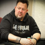 Ein Mann spielt Poker in einem eleganten Casino. Er hält Karten in der Hand und betrachtet konzentriert den Tisch. Um ihn herum sitzen andere Spieler, die ebenfalls Karten halten. Die Szene str