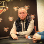 Ein Mann und eine Frau sitzen an einem Pokertisch und spielen Karten. Der Mann hat eine Hand mit Chips vor sich, während die Frau konzentriert ihre Karten betrachtet. Im Hintergrund sind weitere Personen und ein