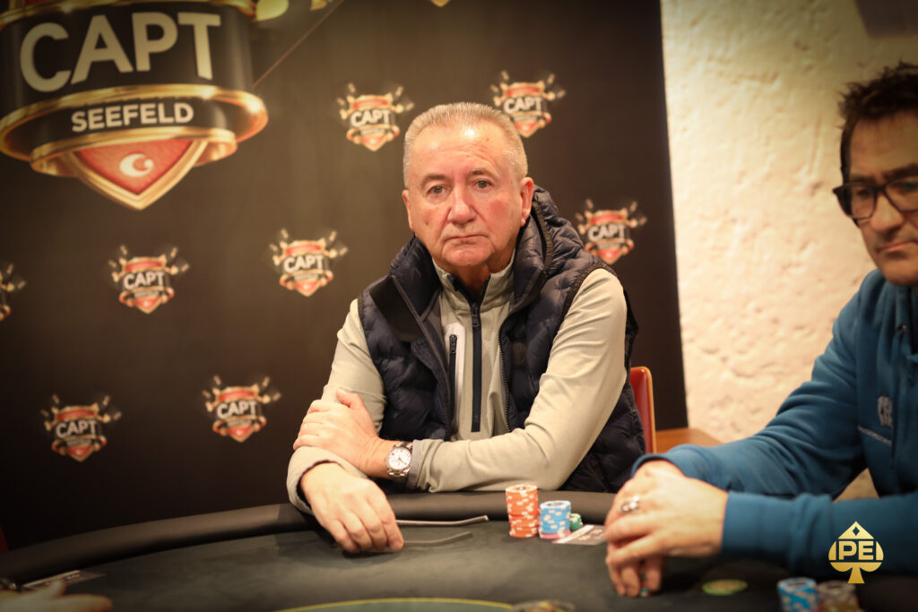 Ein Mann sitzt an einem Pokertisch und betrachtet konzentriert seine Karten. Im Hintergrund sind andere Spieler und Pokerchips zu sehen.