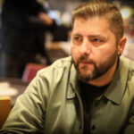 Ein Mann sitzt an einem Pokertisch mit einem Stapel Chips vor sich. Er hält zwei Karten in der Hand und schaut konzentriert auf den Tisch. Im Hintergrund sind andere Spieler und ein Dealer zu sehen
