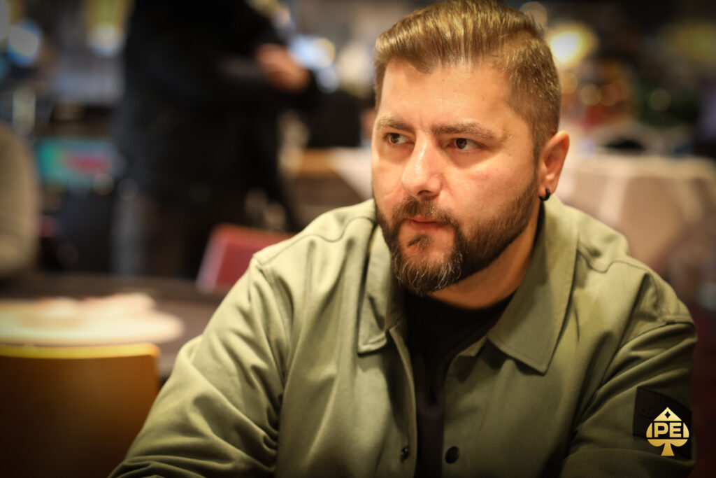 Ein Mann sitzt an einem Pokertisch und hält zwei Spielkarten in der Hand. Er hat eine konzentrierte Miene und betrachtet seine Karten genau. Im Hintergrund sind Pokerchips und andere Spieler zu