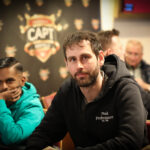 Ein Mann in einem dunkelblauen Anzug und einer blauen Krawatte sitzt an einem Pokertisch. Er hält seine Karten in der Hand und schaut konzentriert auf den Tisch. Im Hintergrund