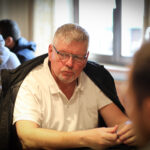 Ein Mann sitzt an einem Pokertisch und hält zwei Karten in der Hand. Er schaut konzentriert auf die Gemeinschaftskarten in der Mitte des Tisches. Im Hintergrund sind andere Spieler und Poker