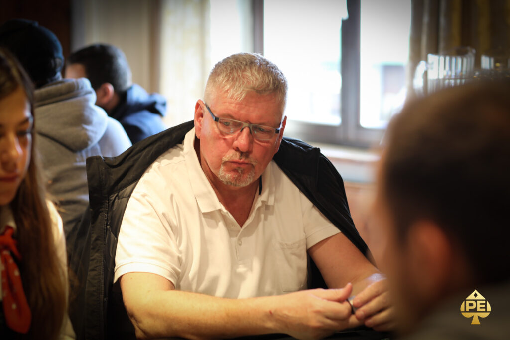 Ein Mann und eine Frau sitzen an einem Pokertisch, konzentriert auf ihre Karten und Jetons. Der Mann hat eine Kappe auf und die Frau trägt eine Brille. Im Hintergrund sind weitere Pokerspi