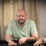 Ein Mann und eine Frau sitzen an einem Pokertisch im Casino und spielen konzentriert. Die Frau hält Karten in der Hand und der Mann schaut auf seine Chips. Im Hintergrund sind andere Spieler und Dealer zu sehen