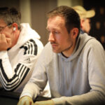 Ein Mann in einem schwarzen Anzug und einer roten Krawatte sitzt an einem Pokertisch und hält zwei Spielkarten in den Händen. Er schaut konzentriert auf den Tisch, umgeben von