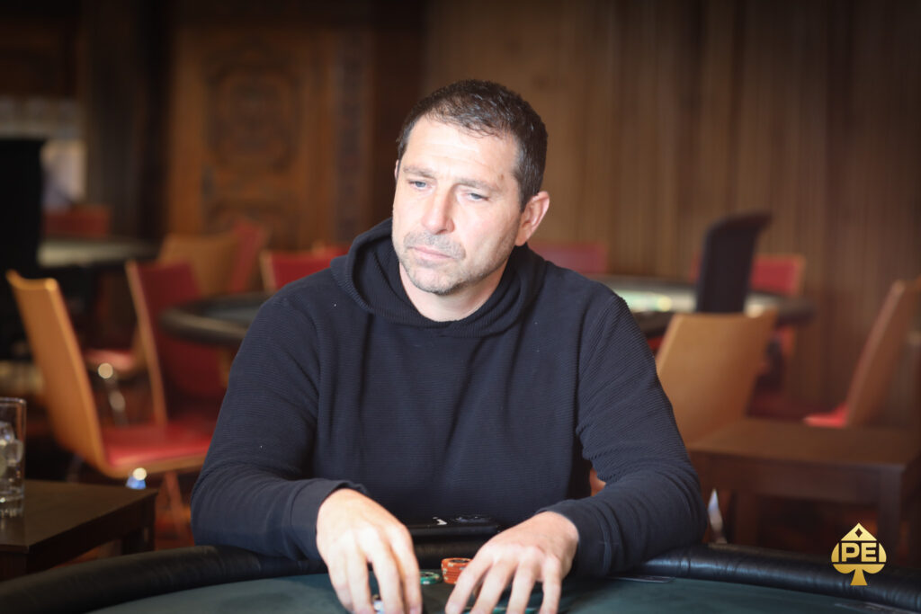 Ein Mann sitzt an einem Pokertisch und hält seine Karten in der Hand, während er konzentriert auf den Tisch schaut. Andere Spieler sind im Hintergrund zu sehen.