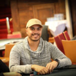 Ein Mann und eine Frau sitzen an einem Pokertisch, sie halten Karten in der Hand und schauen konzentriert auf den Tisch. Im Hintergrund sind Casino-Chips und Getränke zu sehen.