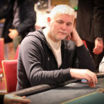 Ein Mann mit Pokerkarten in der Hand, der konzentriert auf seinen Gegner schaut. Im Hintergrund sind Pokerchips und Gläser zu sehen.