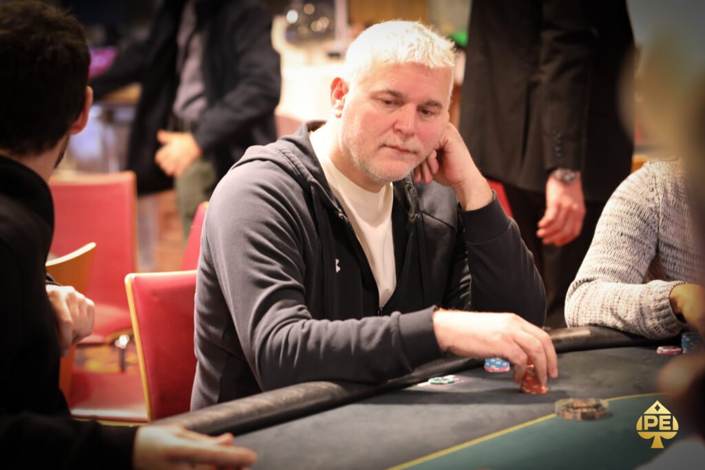 Ein Mann in einem schwarzen Anzug und einer roten Krawatte sitzt an einem Pokertisch und hält seine Karten in der Hand, während er konzentriert auf den Tisch schaut. Um ihn herum sit