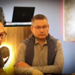 Ein Mann sitzt an einem Pokertisch und hält zwei Spielkarten in der Hand, während er konzentriert auf seine Gegner schaut. Im Hintergrund sind Pokerchips und Getränke zu sehen.