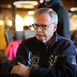 Ein Spieler, der eine Pokerhand hält und seine Karten betrachtet, während er an einem Pokertisch sitzt. Andere Spieler und Chips sind im Hintergrund zu sehen.