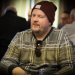 Ein Mann sitzt an einem Pokertisch und hält zwei Spielkarten in der Hand, während er konzentriert auf die Gemeinschaftskarten in der Mitte des Tisches schaut. Im Hintergrund sind weitere Spieler