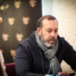 Ein Mann sitzt an einem Pokertisch und hält seine Karten in der Hand, während er konzentriert auf den Tisch schaut. Andere Spieler und Chips sind im Hintergrund zu sehen.