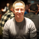 Ein Mann mit einem Pokerchip-Stapel vor sich sitzt an einem Pokertisch, umgeben von anderen Spielern. Er betrachtet seine Karten mit ernstem Gesichtsausdruck.