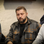 Ein Mann sitzt an einem Pokertisch und hält Karten in der Hand. Er wirkt konzentriert und ist von anderen Pokerspielern umgeben. Im Hintergrund ist eine beleuchtete Decke zu sehen.