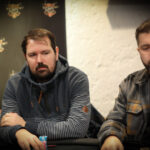 Ein professioneller Pokerspieler mit Sonnenbrille und Kappe, der konzentriert auf seine Karten schaut. Er sitzt an einem grünen Pokertisch mit Chips und anderen Spielern im Hintergrund.