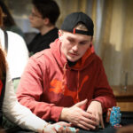 Ein Mann spielt Poker an einem Tisch in einem eleganten Casino. Er hat eine ernste Miene und hält seine Karten in der Hand, während er aufmerksam auf den Tisch schaut. Im Hintergrund sind andere