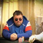 Ein Mann sitzt an einem Pokertisch, hält Karten und betrachtet sie konzentriert, während er von anderen Spielern umgeben ist.