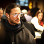 Ein Mann in einem schwarzen Anzug und einer blauen Krawatte spielt im Casino an einem Pokertisch. Er hält Karten in der Hand und schaut konzentriert auf seine Gegner. Die Atmosphäre ist