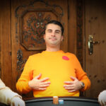 Ein professioneller Pokerspieler mit Sonnenbrille und Kappe sitzt an einem Pokertisch und betrachtet seine Karten mit ernstem Gesichtsausdruck. Im Hintergrund sind andere Spieler und Jetons zu