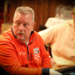 Ein Mann und eine Frau sitzen an einem Pokertisch und spielen eine Partie Poker. Der Mann hält Karten in der Hand, während die Frau konzentriert auf ihre eigenen Karten schaut. Im Hintergrund sind Poker