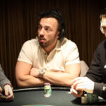 Ein Mann sitzt an einem Pokertisch mit Chips und Karten vor sich. Er hat eine ernste Miene und scheint konzentriert zu sein. Die Umgebung ist elegant und gut beleuchtet.
