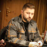 Ein Mann sitzt an einem Pokertisch und betrachtet konzentriert seine Karten. Im Hintergrund sind andere Spieler und Pokerchips zu sehen.