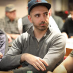 Ein Mann sitzt in einem eleganten Pokerraum an einem Tisch und hält seine Spielkarten in der Hand. Im Hintergrund sind weitere Spieler und eine dekorative Deckenbeleuchtung zu sehen.