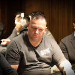 Ein Mann sitzt an einem Pokertisch und hält Karten in der Hand. Er betrachtet konzentriert seine Karten, während andere Spieler im Hintergrund zu sehen sind. Die Atmosphäre ist spannungsgel