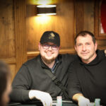Ein Mann sitzt an einem Pokertisch und hält zwei Spielkarten in der Hand, während er konzentriert auf seine Gegner schaut. Die Szene spielt sich in einem eleganten Pokerraum mit grünen