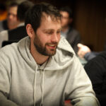 Ein Mann sitzt an einem Pokertisch und hält zwei Spielkarten in der Hand, während er konzentriert auf die Mitte des Tisches schaut. Andere Spieler sind im Hintergrund zu sehen.
