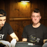 Ein Mann sitzt an einem Pokertisch und hält zwei Karten in der Hand. Er schaut konzentriert auf den Tisch, auf dem Pokerchips und Spielkarten liegen. Im Hintergrund sind andere Spieler