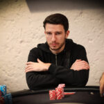 Ein Gruppenfoto von vier Personen, die an einem Pokertisch sitzen und Karten spielen. Sie sind konzentriert und haben Pokerchips vor sich liegen. Die Personen tragen elegante Kleidung und haben ernste