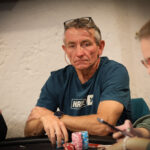 Ein Spieler hält zwei Spielkarten in der Hand, während er konzentriert auf den Pokertisch schaut, um seine nächsten Züge zu planen. Im Hintergrund sind Pokerchips und andere Spieler zu sehen.