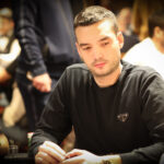 Ein Mann sitzt an einem Pokertisch, umgeben von anderen Spielern. Er hält Karten in der Hand und schaut konzentriert auf den Tisch. Die Szene spielt sich in einem eleganten Pokerraum