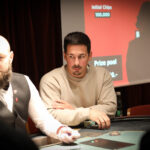 Ein Mann mit Bart und Brille sitzt an einem Pokertisch und hält Karten in der Hand. Er wirkt konzentriert und schaut ernst in die Kamera. Hinter ihm ist eine verschwommene Gl