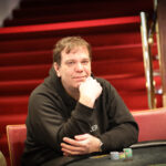 Ein Mann in einem schwarzen Anzug spielt Poker an einem eleganten Tisch in einem luxuriösen Casino. Er hält Karten in der Hand und hat einen konzentrierten Blick. Im Hintergrund sind andere Spieler und